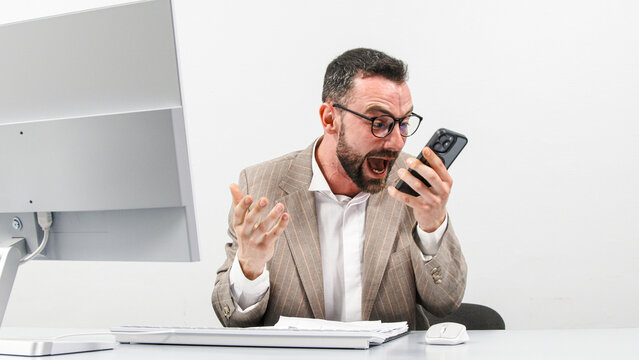 Angry businessman shouting and gesticulating while talking on cell phone in front of computer display on light background. The concept of rage, aggression and discontent.