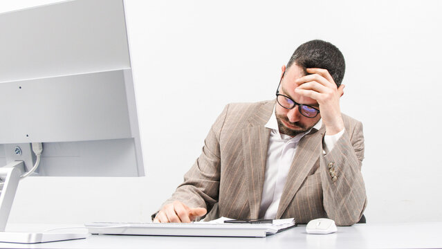 Tired, focused business man working at a computer in a bright office. The man looks frustrated while working. The concept of stress, frustration and burnout in the workplace.