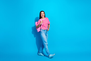 Stylish young woman wearing a pink striped t-shirt, jeans, and backpack in front of a vibrant blue...