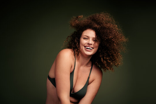 Smiling young woman with curly hair in green lingerie posing confidently with radiant charm and natural beauty on a clean studio background