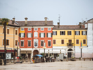 Obraz premium A rainy day in the historic town square of Palmanova, Italy, featuring old buildings with colorful facades, statues, and palm trees under a cloudy sky.