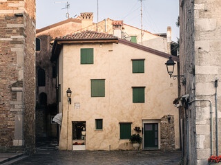 A picturesque historic house in Grado, Italy, with warm beige walls, green shutters and a rustic tiled roof. The narrow stone-paved street and vintage lanterns enhance the old European town atmosphere