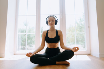 A peaceful young woman sits cross-legged, meditating while wearing headphones in a serene, sunlit...