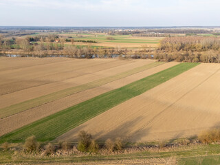 Obraz premium Aerial view of a vast agricultural landscape featuring striped fields, a river, and scattered trees under a clear sky in early spring.