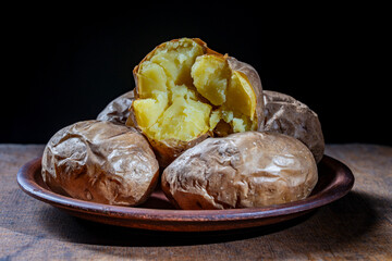 Baked potatoes whole in their skins on a clay plate, closeup. Simple Ukrainian food on black background. Jacket potato for eat