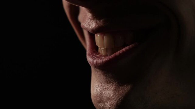 Close-up of a sinister smile in dramatic low lighting, showing slightly parted lips and visible teeth, evoking a sense of mystery, tension, and unease in a dark. The man gloats. Scary wide smile