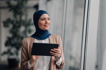Smiling muslim businesswoman holding tablet and looking out office window