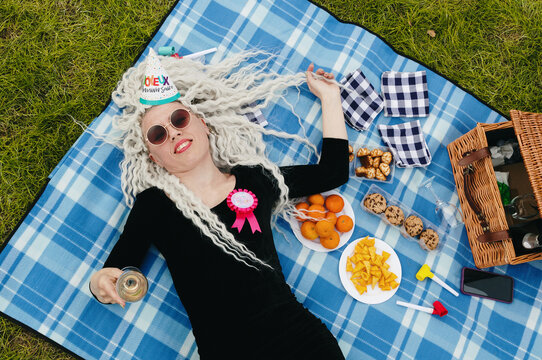 Portrait of blonde in birthday hat with glass of champagne in hand on picnic in park.