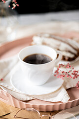 White cup filled with steaming coffee on a saucer, placed on a pink tray. Beside it lies a French eclair with cream. A small sprig of pink flowers adds decoration.