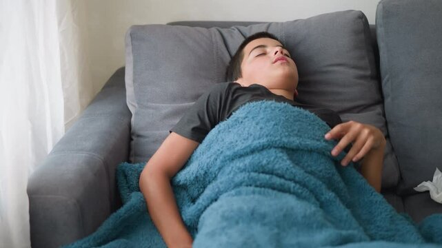 Uncomfortable child sleeping covered in blanket adjusting on couch for comfort with tissue nearby, showing signs of discomfort during rest in peaceful indoor setting