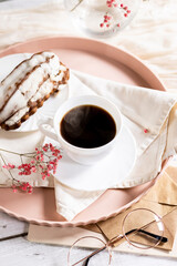 White cup filled with steaming coffee on a saucer, placed on a pink tray. Beside it lies a French eclair with cream. A small sprig of pink flowers adds decoration.