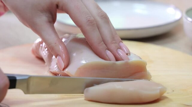 Close-up of hands slicing raw chicken breast on a wooden cutting board with a sharp knife, focusing on clean preparation and kitchen hygiene essentials