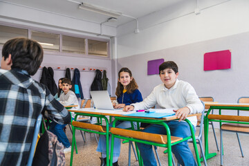 Obraz premium Elementary school students sitting at desks using laptop computer and learning together in classroom