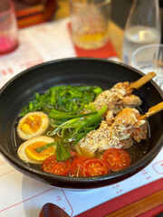 Ramen bowl with vegetables, egg and chicken skewer on wooden table in restaurant. Comfort food and cultural dining experience