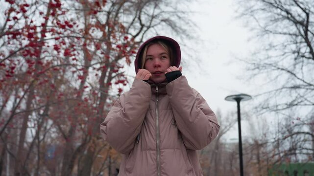 Woman bundled in warm winter clothing adjusts her hoodie while walking through a snowy park, she holds her hands near her face to keep warm, surrounded by bare trees with red berries