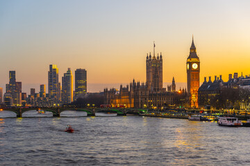 Fototapeta premium At dusk, the River Thames reflects the warm glow of evening as Big Ben and Westminster Palace stand majestically in the background. A boat glides gently on the water, creating a serene atmosphere.