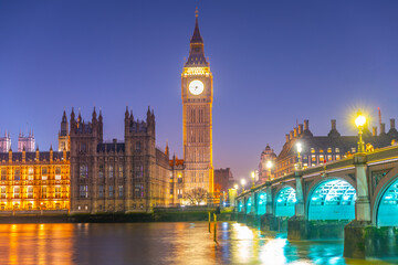 Fototapeta premium The iconic Houses of Parliament and Big Ben stand majestically against the night sky, brilliantly illuminated. Westminster Bridge glows with lights reflecting on the calm river.