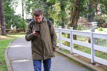 Man wearing a green jacket and backpack walks along a park path while intently looking at his...