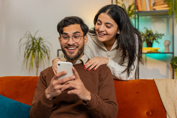 Smiling Indian woman talking with boyfriend swiping smartphone sitting on sofa at home. Multiethnic young couple using mobile phone together making order online delivery in living room apartment.