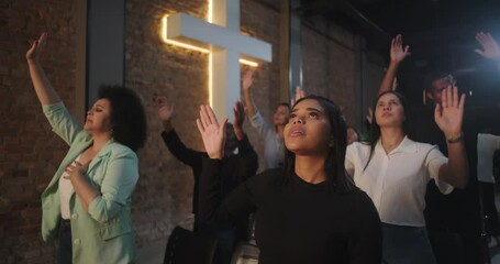 Diverse worshippers raising hands in worship service with glowing cross in background, people deeply involved in prayer and praise