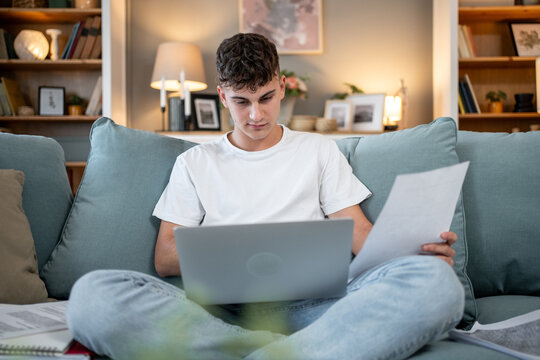 Focused teen studying online and reviewing documents on sofa at home