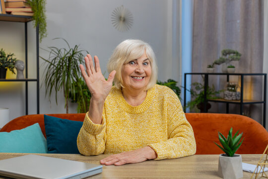 Hello. Senior woman smiling friendly at camera waving hands gesturing hello hi greeting goodbye welcoming with hospitable expression at home indoors. Elderly mature grandmother sits on couch at table