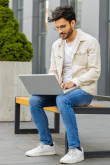 Happy Indian man freelancer working on laptop computer sends messages, makes online purchases, watching movies, reading email outdoors. Arabian guy sitting on street bench. Business people. Vertical