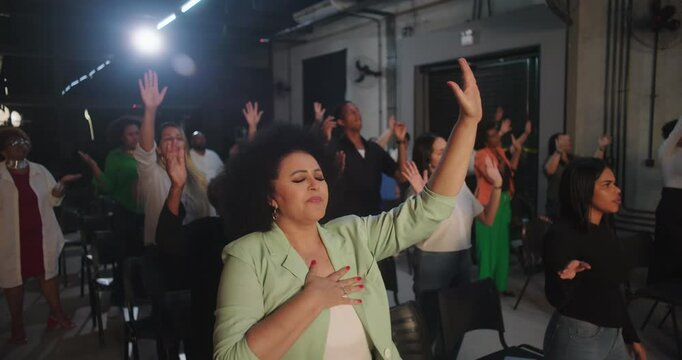 Diverse group in worship session, woman in focus placing hand on chest with raised arm, emotional and spiritual expression, community gathering in indoor setting