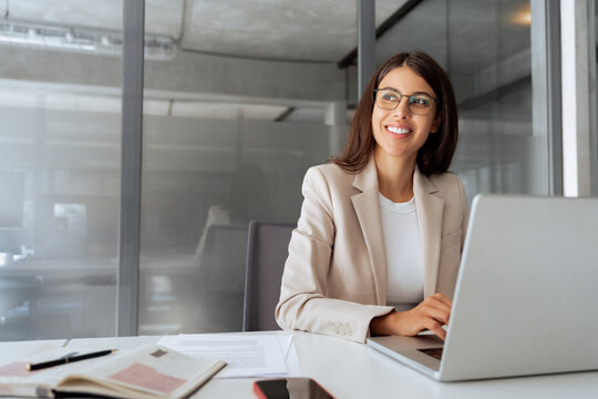 Smiling middle aged business woman using laptop at work in office. Professional female executive entrepreneur in suit working on computer pc at company workplace, looking dreaming aside. Copy space