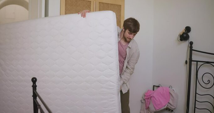 A happy brunette guy in a white jacket brings a mattress with his girlfriend into the new bedroom during a house move
