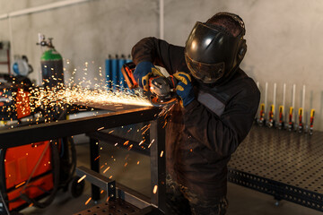 Metalworker wearing protective gear uses an angle grinder, creating a shower of sparks while...