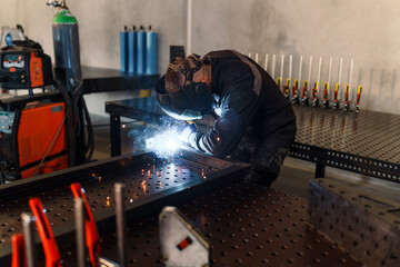 Welder wearing protective gear skillfully joins a metal frame on a welding table, utilizing...