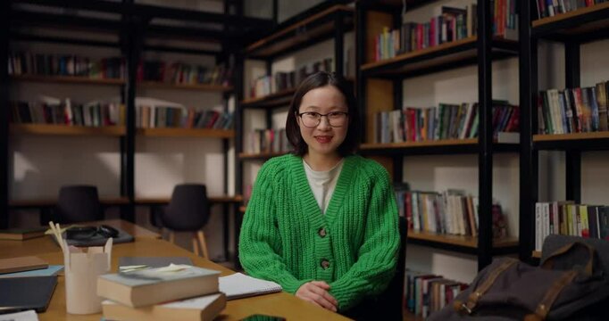 Portrait of a happy Asian girl in glasses and a green sweater who sits at a table in the library