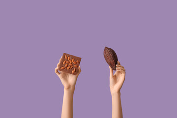 Female hands with sweet chocolate and cocoa pod on lilac background