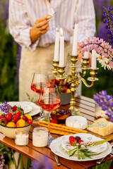 Two glasses of rose wine, flowers in a vase, silverware, fruits. Sunset, golden hour, atmospheric candlelight. Romantic table decor for a loving couple on blooming meadow with purple lupines