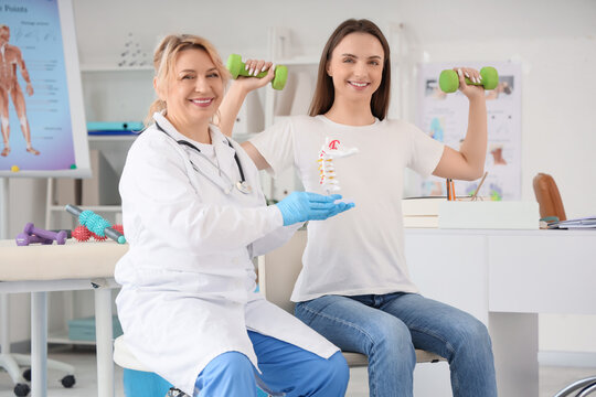 Mature happy female osteopath holding spine model and young woman doing exercises with dumbbells in clinic - Powered by Adobe