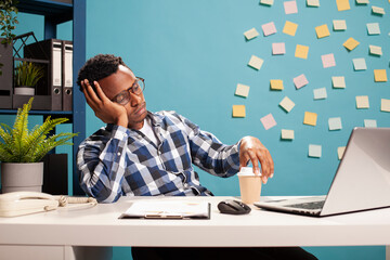 Tired marketing specialist sitting at desk, reviewing key performance indicators on laptop, deep in...