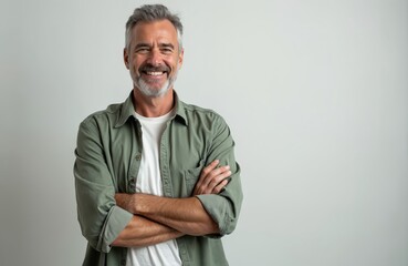 Smiling mature man stands against white background. Attractive caucasian male in casual shirt crosses arms. Smiling senior businessman, looking in camera, happy with positive face expression.