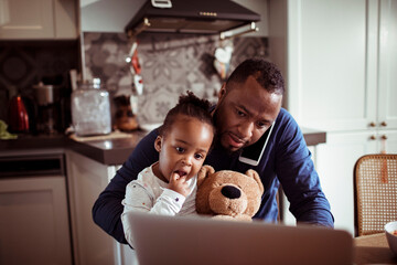 Single father multitasking talking on the phone and working on his laptop while having breakfast with his baby daughter in the kitchen