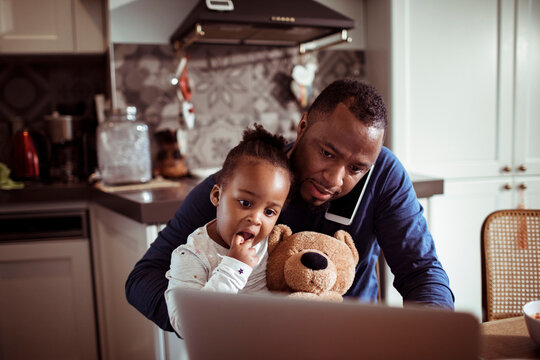 Single father multitasking talking on the phone and working on his laptop while having breakfast with his baby daughter in the kitchen