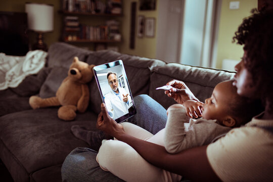 Worried mother talking to her daughters pediatrician over a video call on her tablet while holding her daughter on a couch in the living room