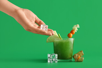 Female hand with ice cube and glass of green bloody mary cocktail garnished with physalis berries...