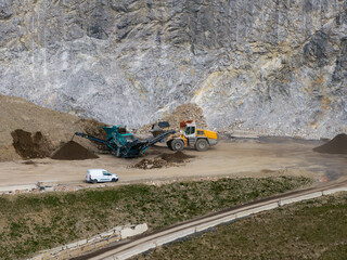 A yellow and white excavator works near a rocky cliff in the Austrian Alps, with a blue industrial machine, a white van, and a narrow paved road nearby.