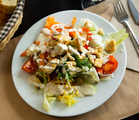 Caesar salad with slices of fried chicken, fresh tomatoes, corn and olives on a white plate. Delicious serving of meal with vegetables and meat. Lunch option in a cafe