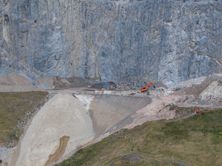 An orange excavator operates at a construction site in the Austrian Alps, surrounded by rocky...