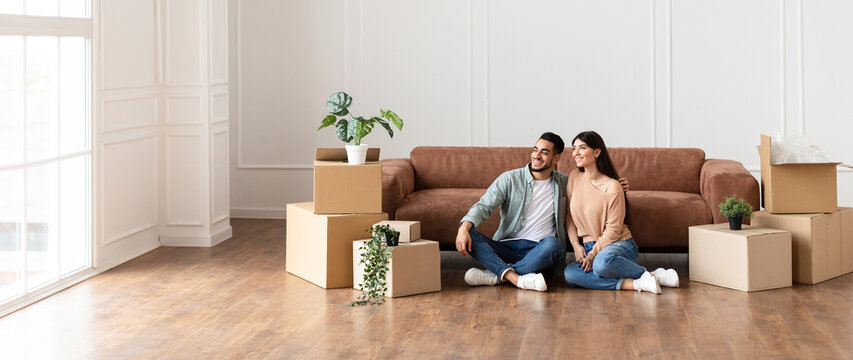 Moving Day. Lovely couple sitting on the floor in new home with cardboard boxes around. Happy man and woman dreaming, looking aside at window, free copy space, panoramic banner for advertisement