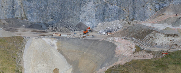 An orange excavator operates on a rocky construction site in the Austrian Alps, surrounded by steep...