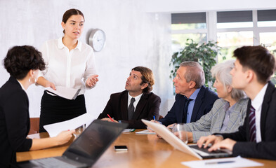 Positive young businesswoman discussing new business project with members of team gathered around table in office meeting room
