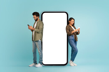 Stylish beautiful young eastern couple posing by huge phone with white blank screen, cheerful handsome indian man and pretty long-haired woman using smartphones, colorful background, mockup