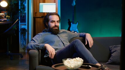 Amused man sitting on couch laughing while watching funny comedy movie, eating popcorn. Cheerful person in apartment consuming snacks and being amused by hilarious joke in film scene, camera A
